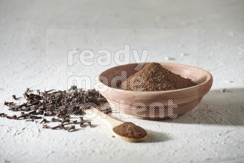 A wooden bowl and wooden spoon full of cloves powder with cloves spread on textured white flooring