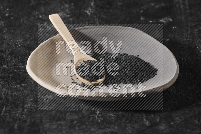 A multicolored pottery plate full of black seeds and wooden spoon full of seeds on a textured black flooring