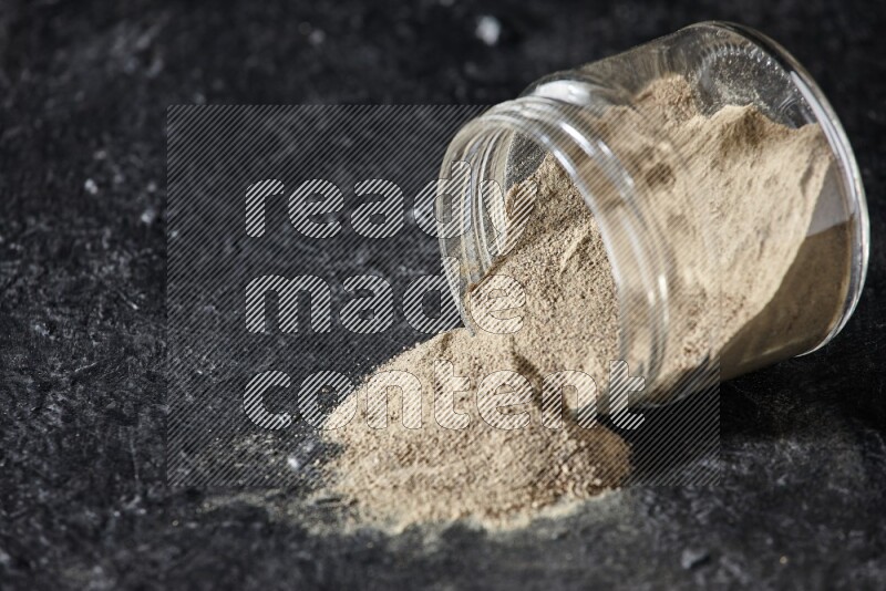 A flipped glass jar full of white pepper powder with spilled powder on textured black flooring