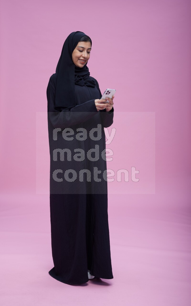 Saudi woman wearing Abaya standing texting on phone on pink background
