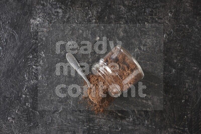 A flipped glass jar and metal spoon full of cloves powder with cloves spread on a textured black flooring