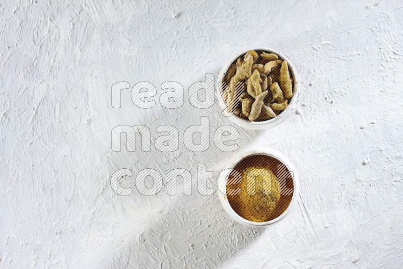 وعاءان فخاريان بيج ممتلئان ببودرة الكركم وأصابع الكركم المجففة على خلفية بيضاء