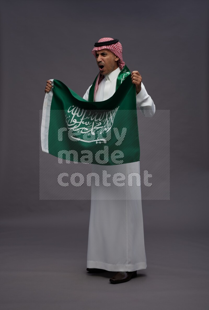 Saudi man wearing thob and shomag standing holding Saudi flag on gray background