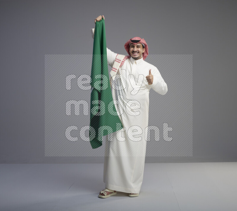 A saudi man standing wearing thob and red shomag holding big saudi flag on gray background