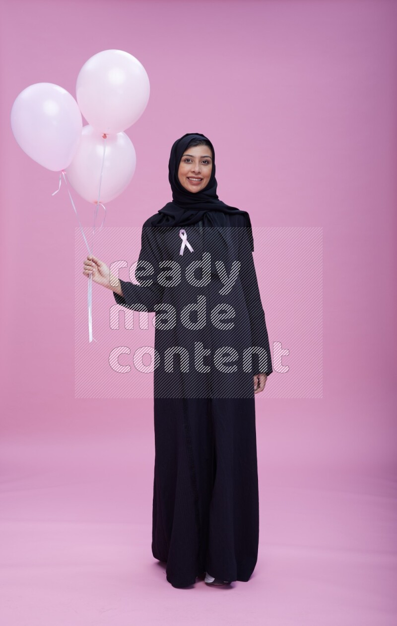 Saudi woman wearing pink ribbon on Abaya standing holding balloons on plain pink background