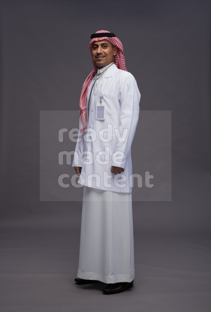 Saudi man wearing thob with lab coat and shomag with pocket employee badge with stethoscope standing interacting with the camera on gray background