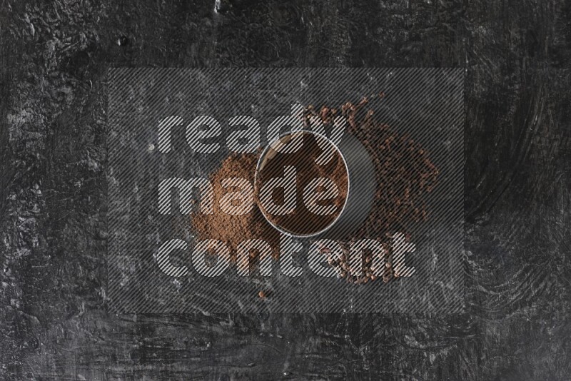 A black pottery bowl full of cloves powder spilled out of it with whole grains on a textured black flooring