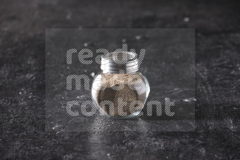 A glass spice jar full of black pepper powder on textured black flooring