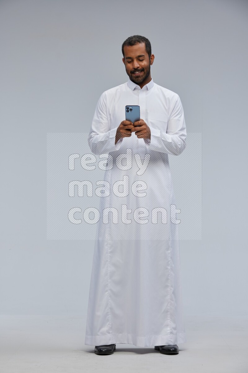 Saudi man wearing Thob standing texting on phone on Gray background