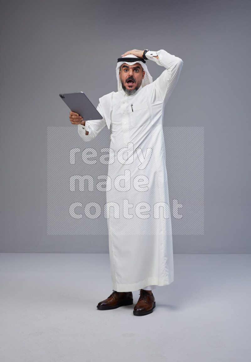 Saudi man Wearing Thob and white Shomag standing working on tablet on Gray background