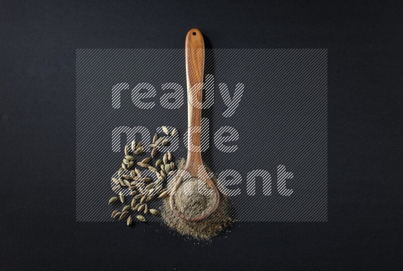 A wooden ladle full of cardamom powder and cardamom seeds beside it on black flooring