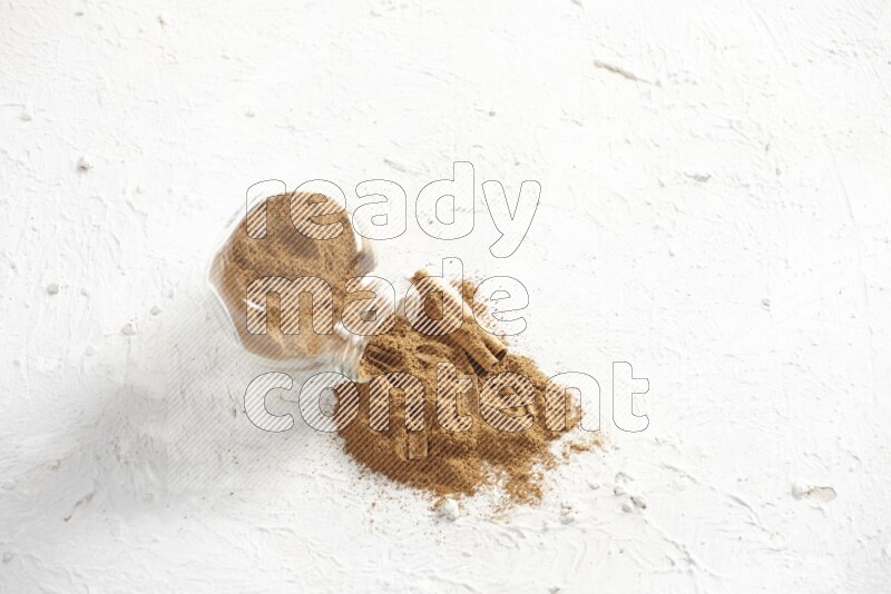Flipped glass jar full of cinnamon powder with some pieces of cinnamon sticks on a textured white background