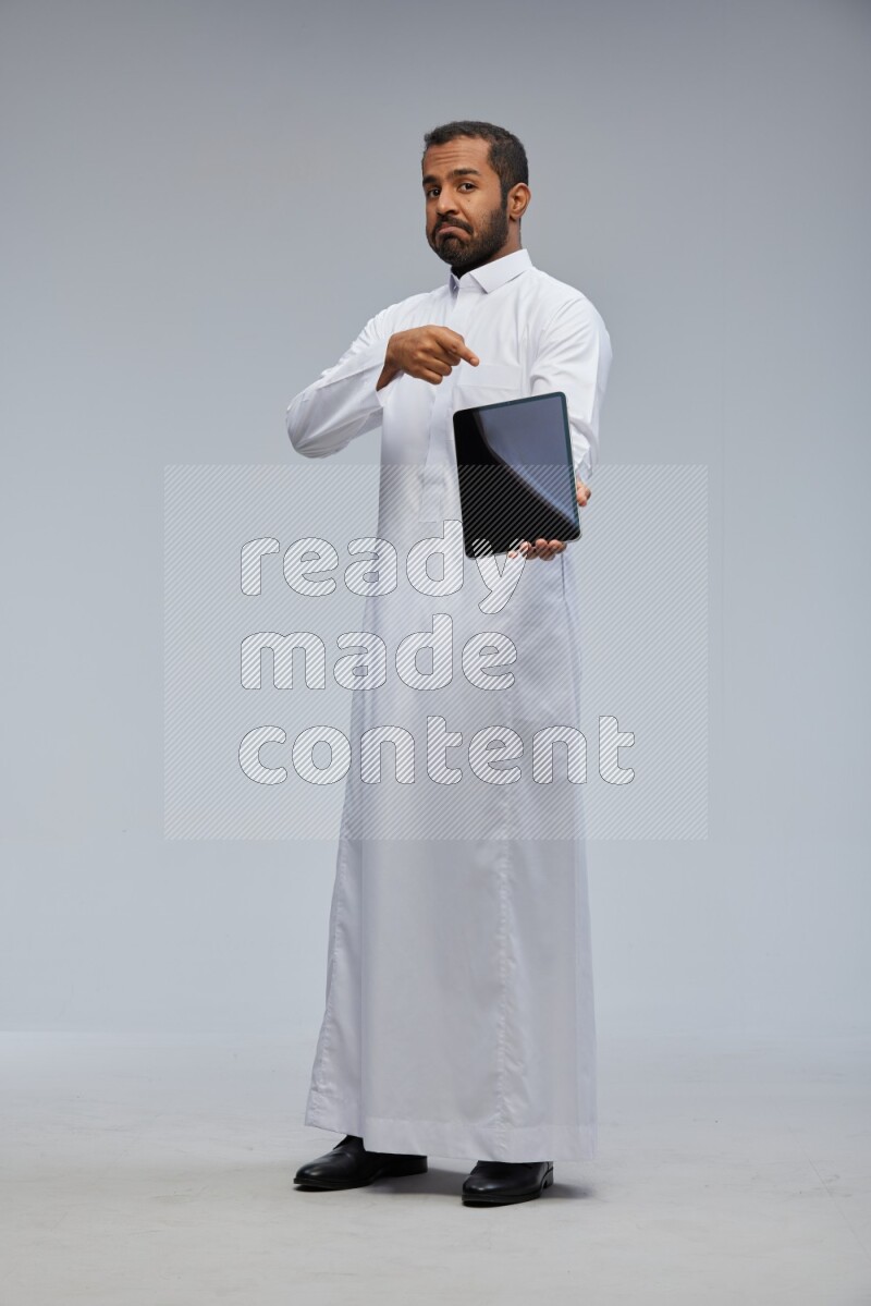 Saudi man wearing Thob standing showing tablet to camera on Gray background