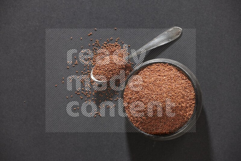 A black pottery bowl full of garden cress seeds and metal spoon full of the seeds on a black flooring