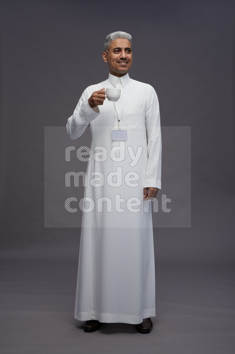 Saudi man wearing thob with neck strap employee badge standing holding mug on gray background