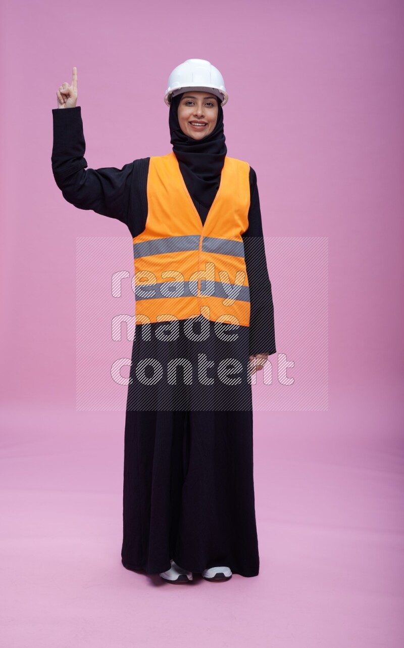 Saudi woman wearing Abaya with engineer vest and helmet standing interacting with the camera on pink background