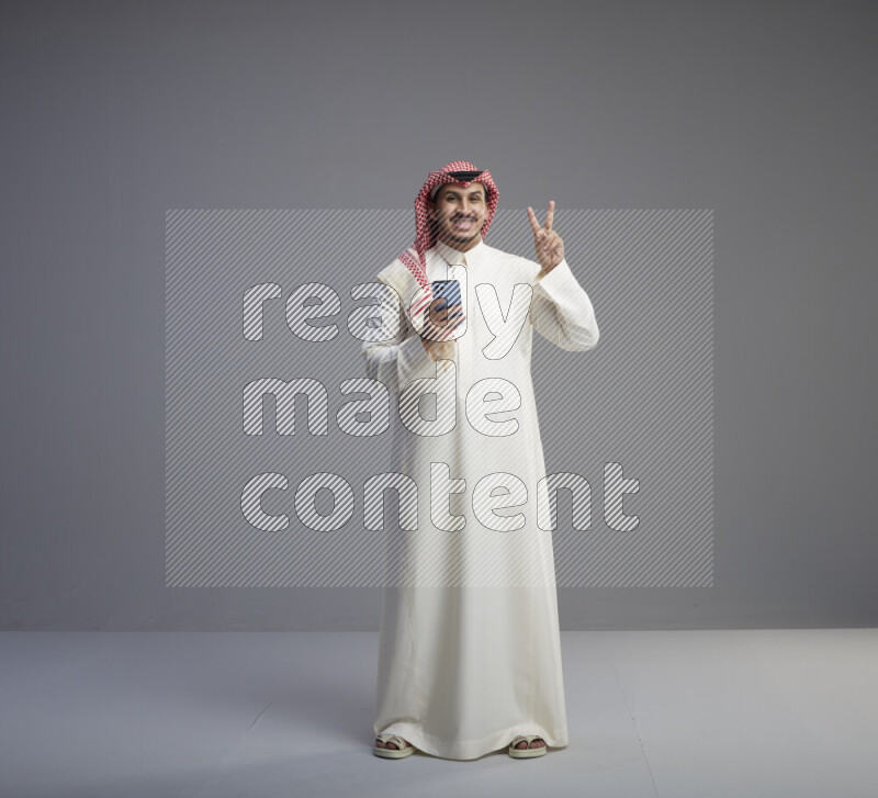 A  Saudi man standing wearing thob and red shomag texting on phone on gray background