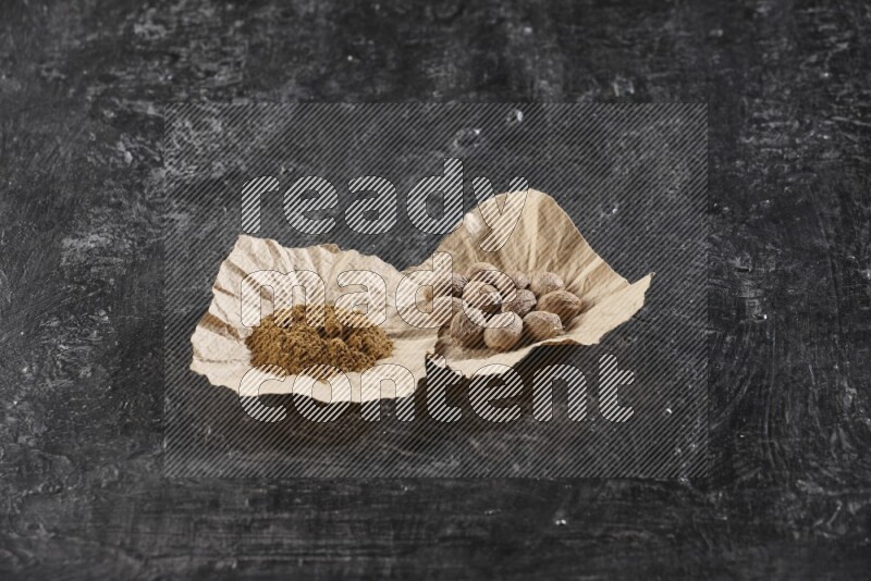 2 crumpled piece of paper full of nutmeg seeds and powder on a textured black flooring