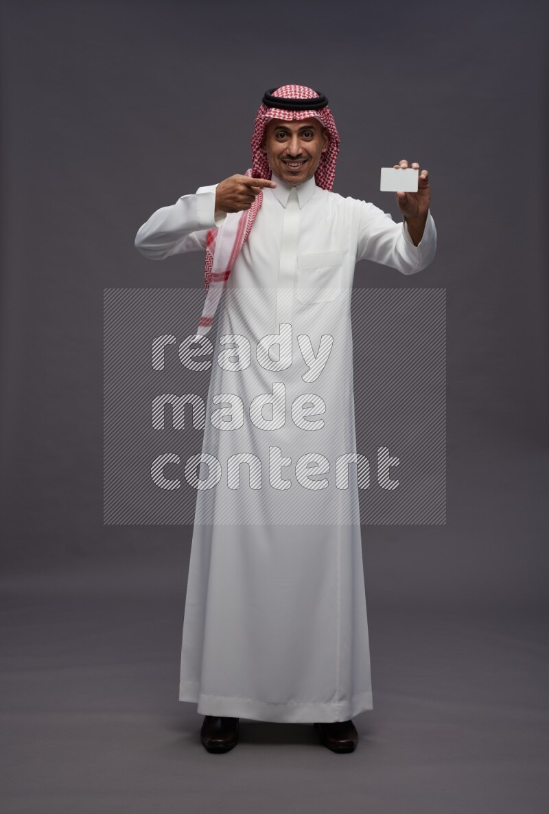 Saudi man wearing thob and shomag standing holding ATM card on gray background