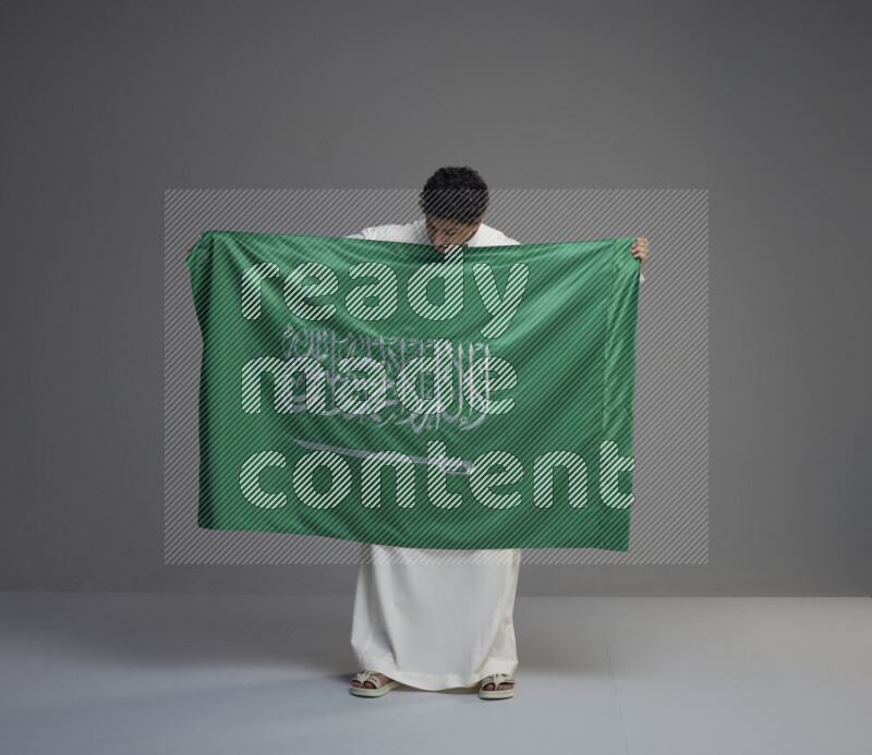 A saudi man standing wearing thob holding big saudi flag on gray background