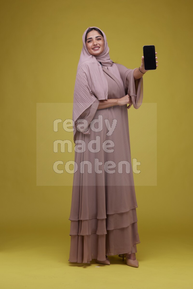 A woman Showing her Phone screen on a Yellow Background wearing Brown Abaya with Hijab