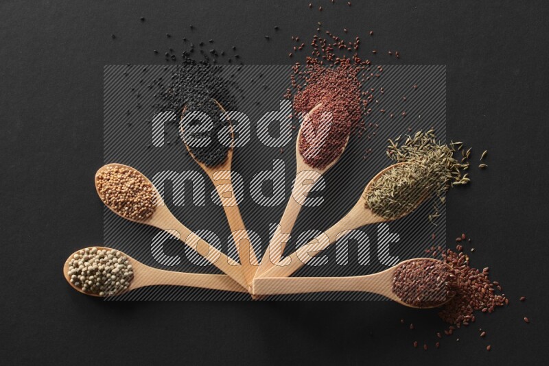 wooden spoons filled with white peppers, mustard seeds, black seeds, garden cress, cumin and flax on black flooring