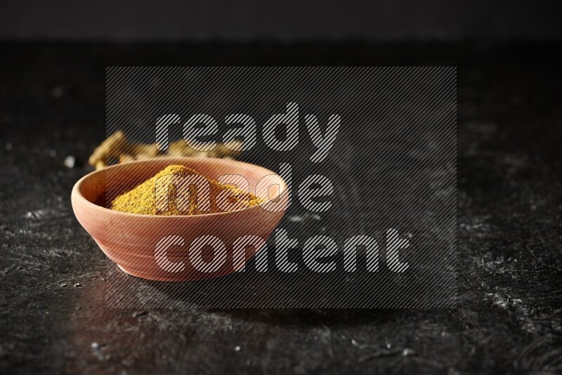 طبق خشبي ممتلئ ببودرة الكركم مع أصابع الكركم المجففة على خلفية سوداء