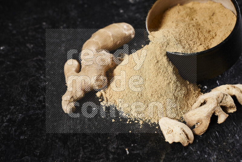 A black pottery bowl full of ground ginger powder with fallen powder from it on black background