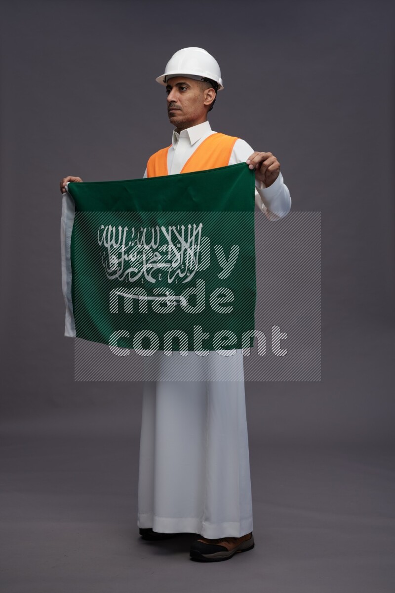 Saudi man wearing thob with engineer vest standing holding Saudi flag on gray background