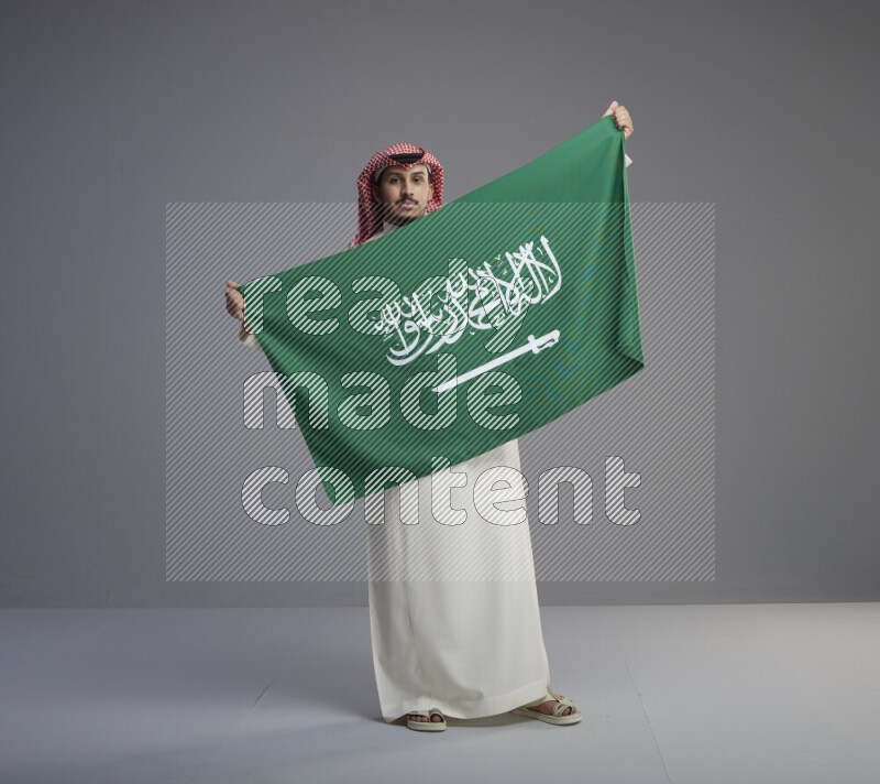 A saudi man standing wearing thob and red shomag holding big saudi flag on gray background