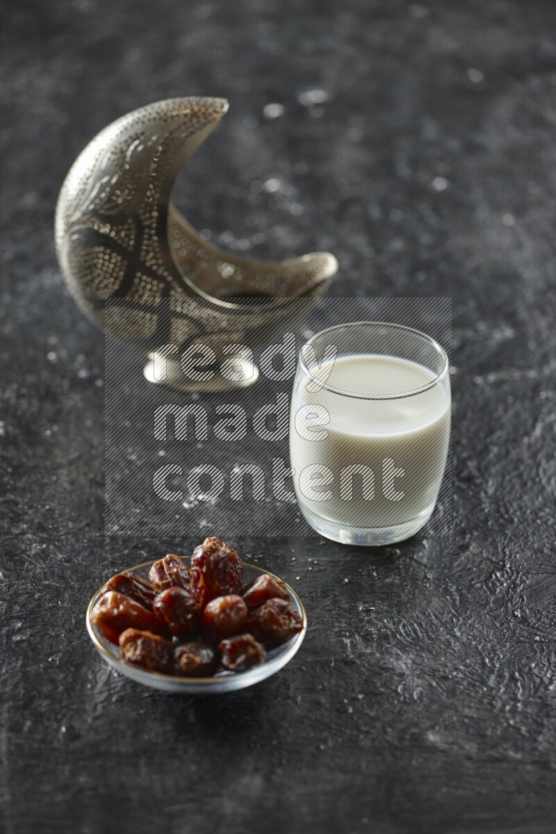 A silver lantern with different drinks, dates, nuts, prayer beads and quran on textured black background
