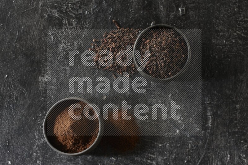 2 Black pottery bowls full of cloves and the other full of cloves powder on textured black flooring