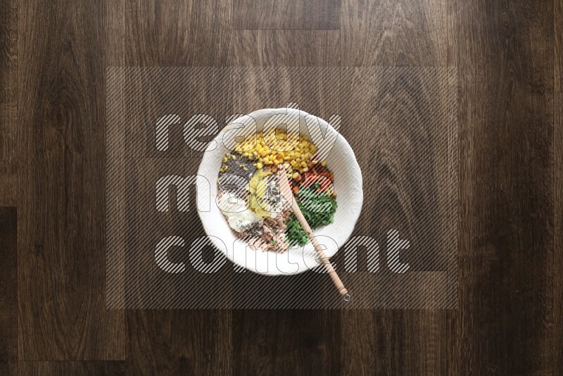 A white bowl full of tuna, colored bell pepper, sour cream, corn, parsley, black pepper powder and sauce, with wooden spoon on wooden background