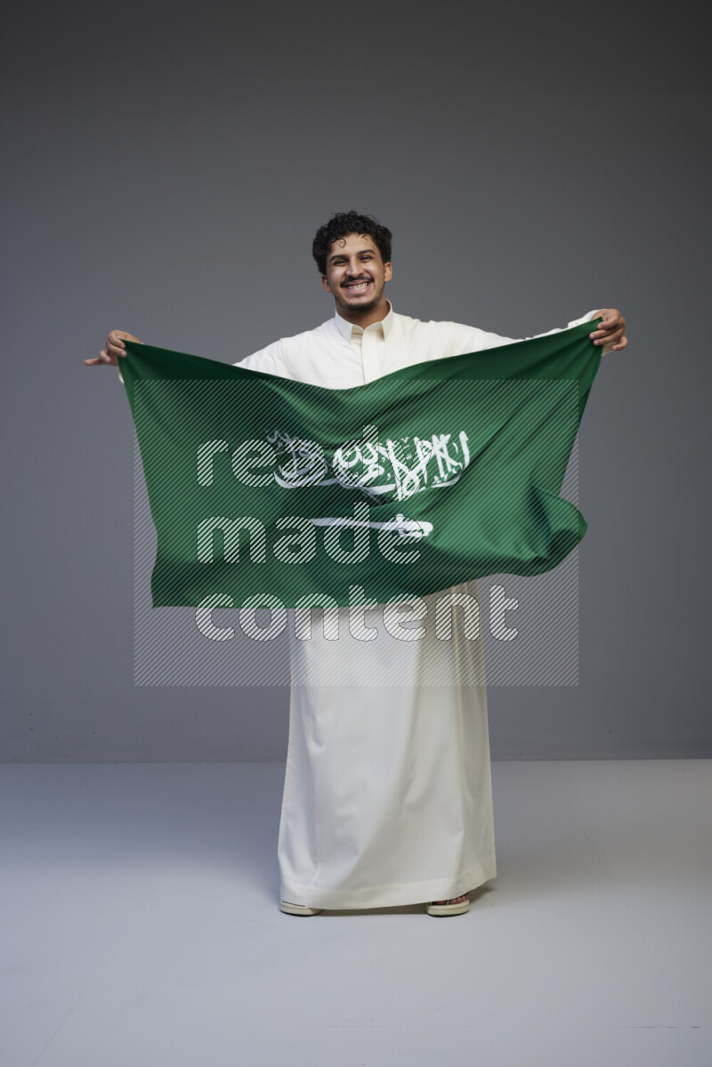 A Saudi man standing wearing thob holding big Saudi flag on gray background