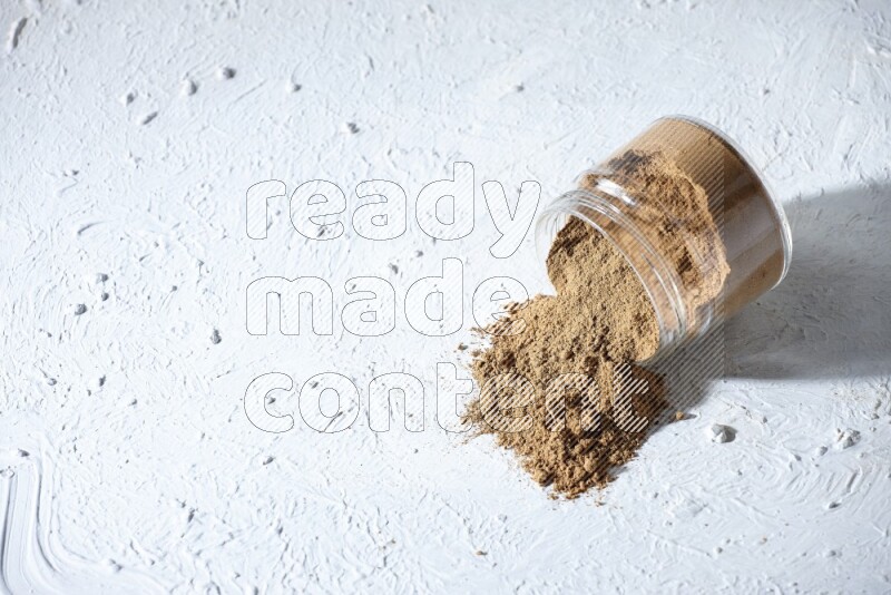 A flipped glass jar full of allspice powder and powder spilled out of it on a textured white flooring