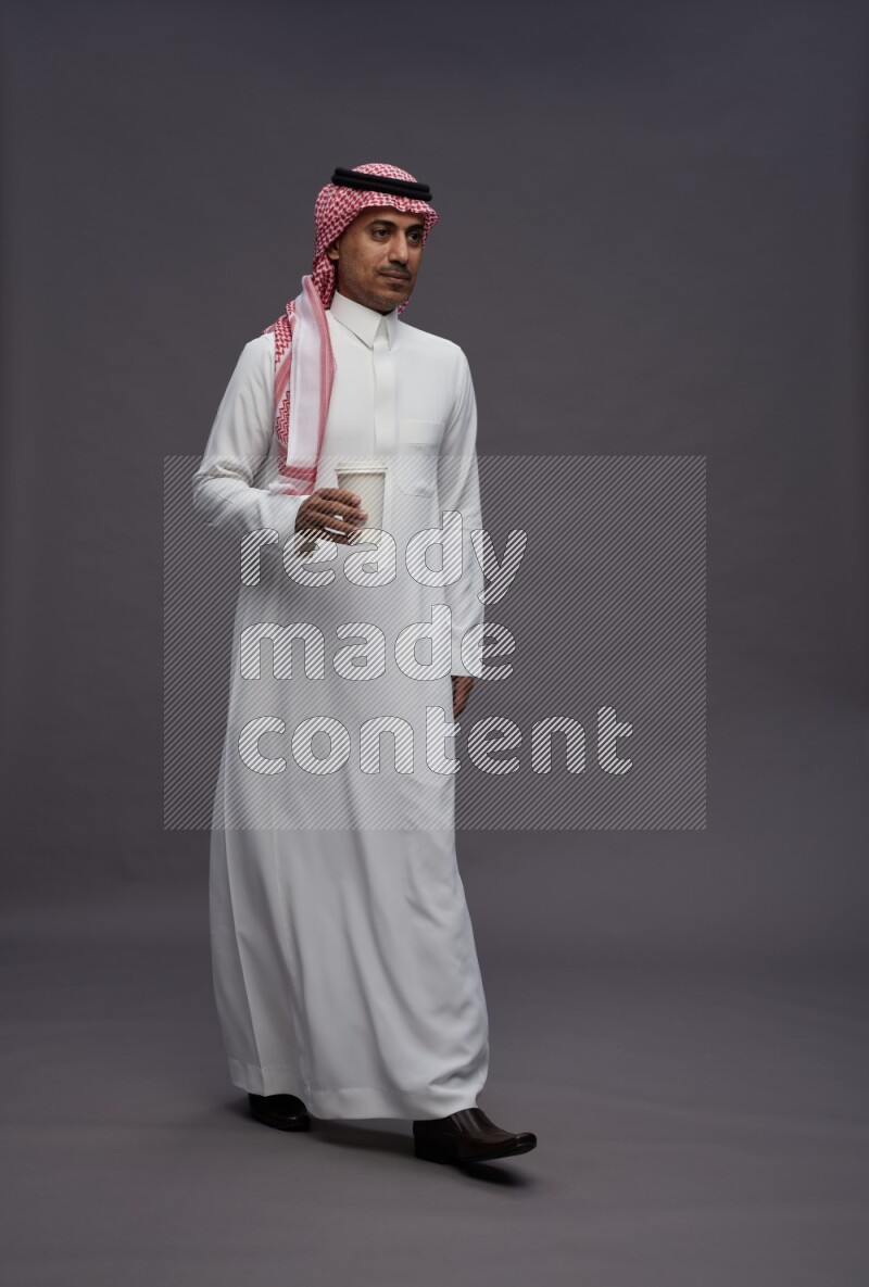 Saudi man wearing thob and shomag standing holding paper cup on gray background