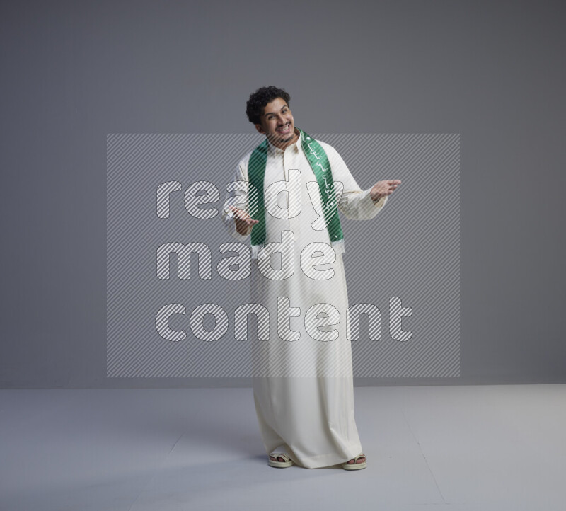 A Saudi man standing wearing thob and Saudi flag scarf on gray background