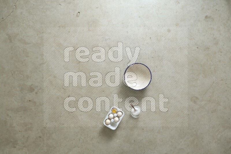 A ceramic bowl beside a carton of eggs and glass bowl full of sugar, vanilla extract, flour on beige marble background