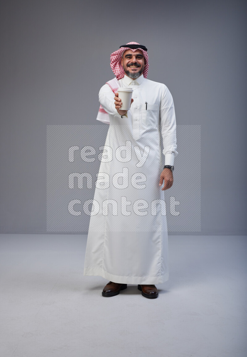 Saudi man Wearing Thob and red Shomag standing holding paper cup on Gray background