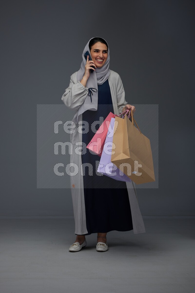 A Saudi woman wearing a light gray Abaya and head scarf standing and holding shopping bags making different poses eye level on a grey background