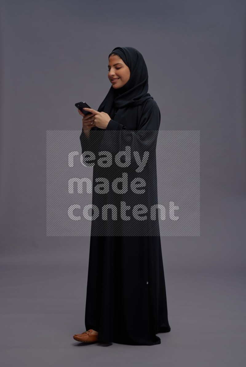 Saudi woman wearing Abaya standing texting on phone on gray background