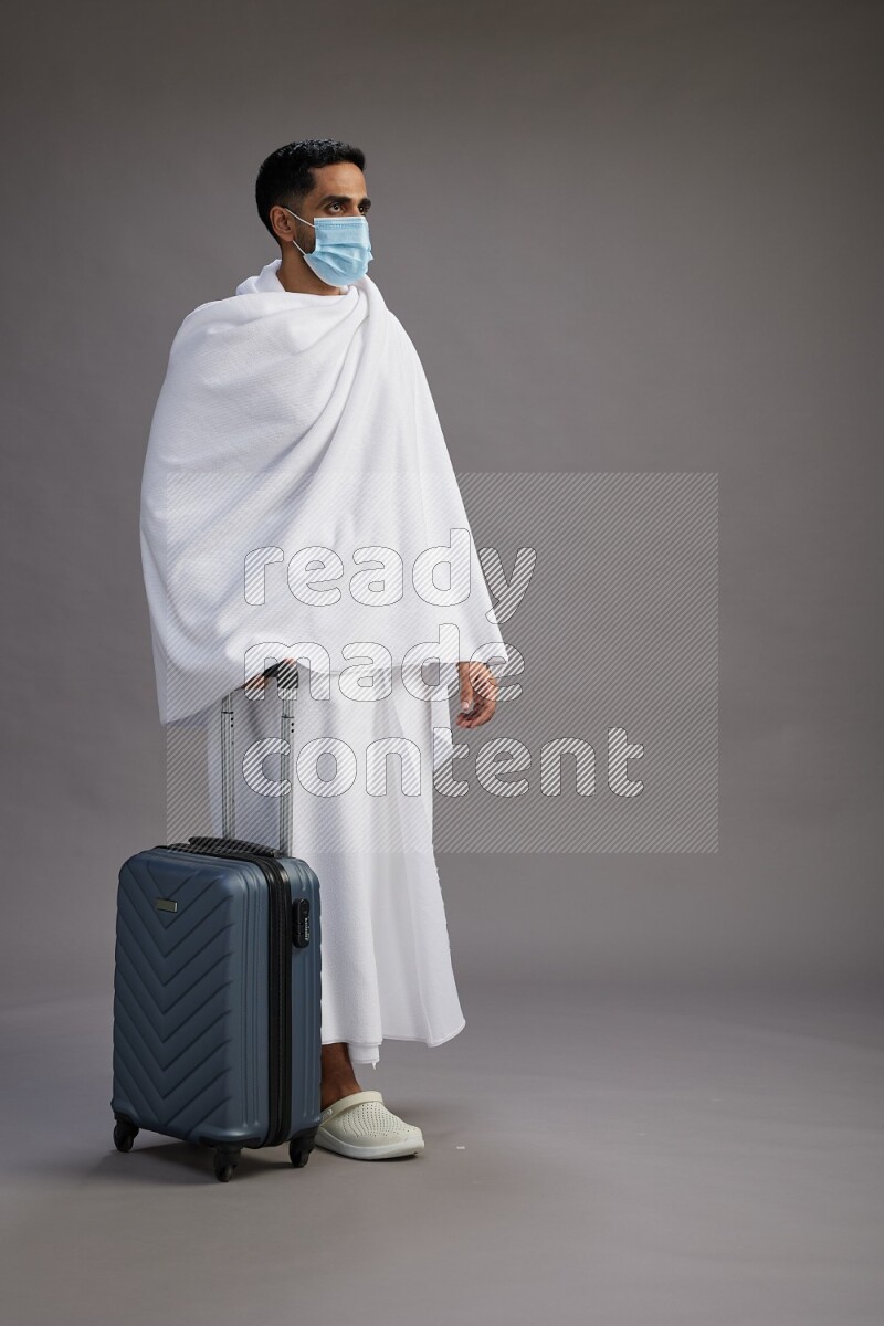 A man wearing Ehram with face mask Standing holding traveling bag on gray background