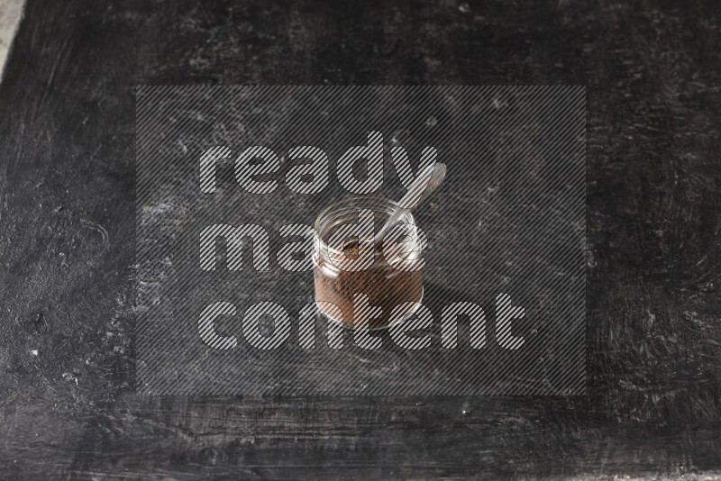 A glass jar full of cloves powder with a metal spoon on a textured black flooring