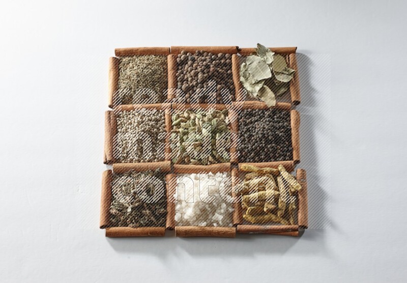 9 squares of cinnamon sticks full of cardamom in the middle surrounded by turmeric, salt, dried basil, white pepper, cumin, allspice, bay laurel leaves and black pepper on white flooring