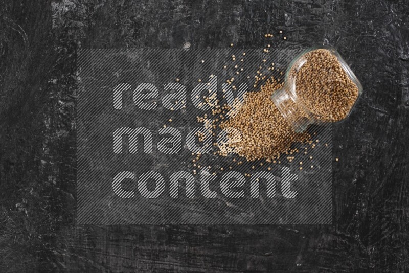 A glass spice jar full of mustard seeds and jar is flipped and seeds spread out on a textured black flooring