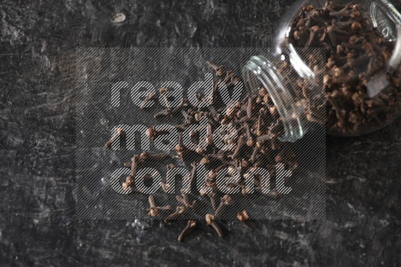 A flipped glass spice jar full of cloves and cloves came out of it on textured black flooring