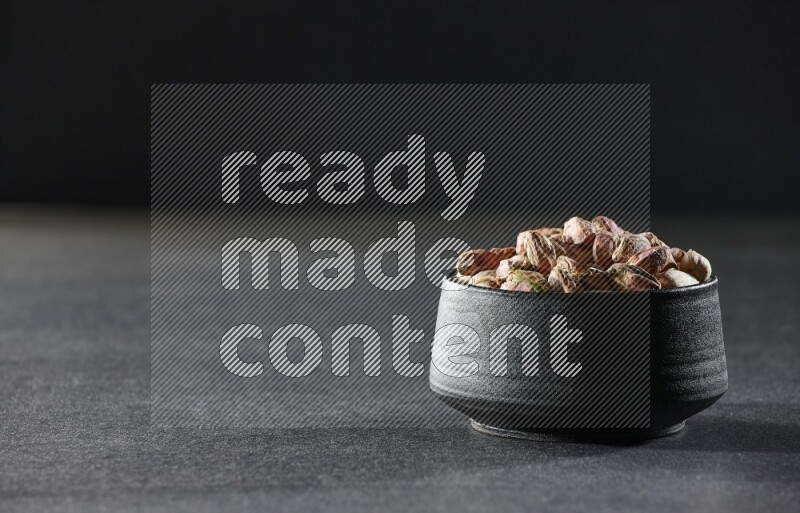 A black pottery bowl full of peeled pistachios on a black background in different angles
