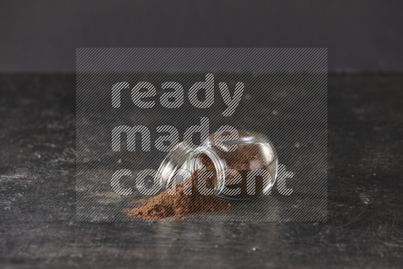 A flipped glass spice jar full of cloves powder and powder came out of it on textured black flooring