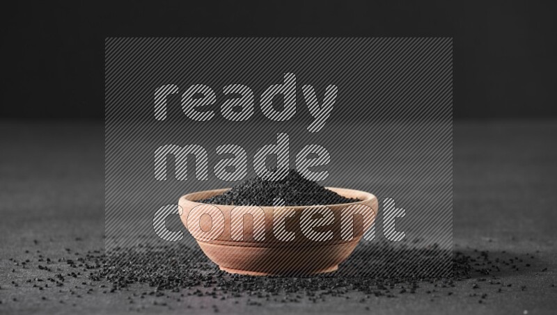 A wooden bowl full of black seeds surrounded by seeds on a black flooring
