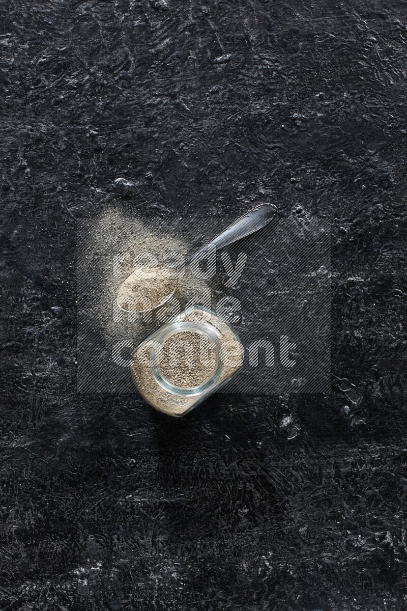 Herbal glass jar and metal spoon full of white pepper powder on textured black flooring